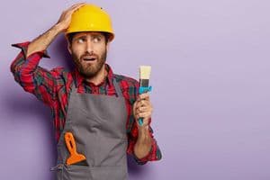Nervous construction worker holds paint brush, touches head and looks aside, wears safety hardhat, dressed in apron, puzzled with having much work, poses on purple wall with blank copy space