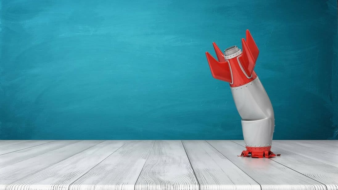 3d rendering of a red and silver realistic model of a retro rocket stands crashed into a wooden desk on a blue background. Failed launch. Technological progress. Technical mistake.