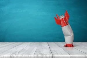 3d rendering of a red and silver realistic model of a retro rocket stands crashed into a wooden desk on a blue background. Failed launch. Technological progress. Technical mistake.